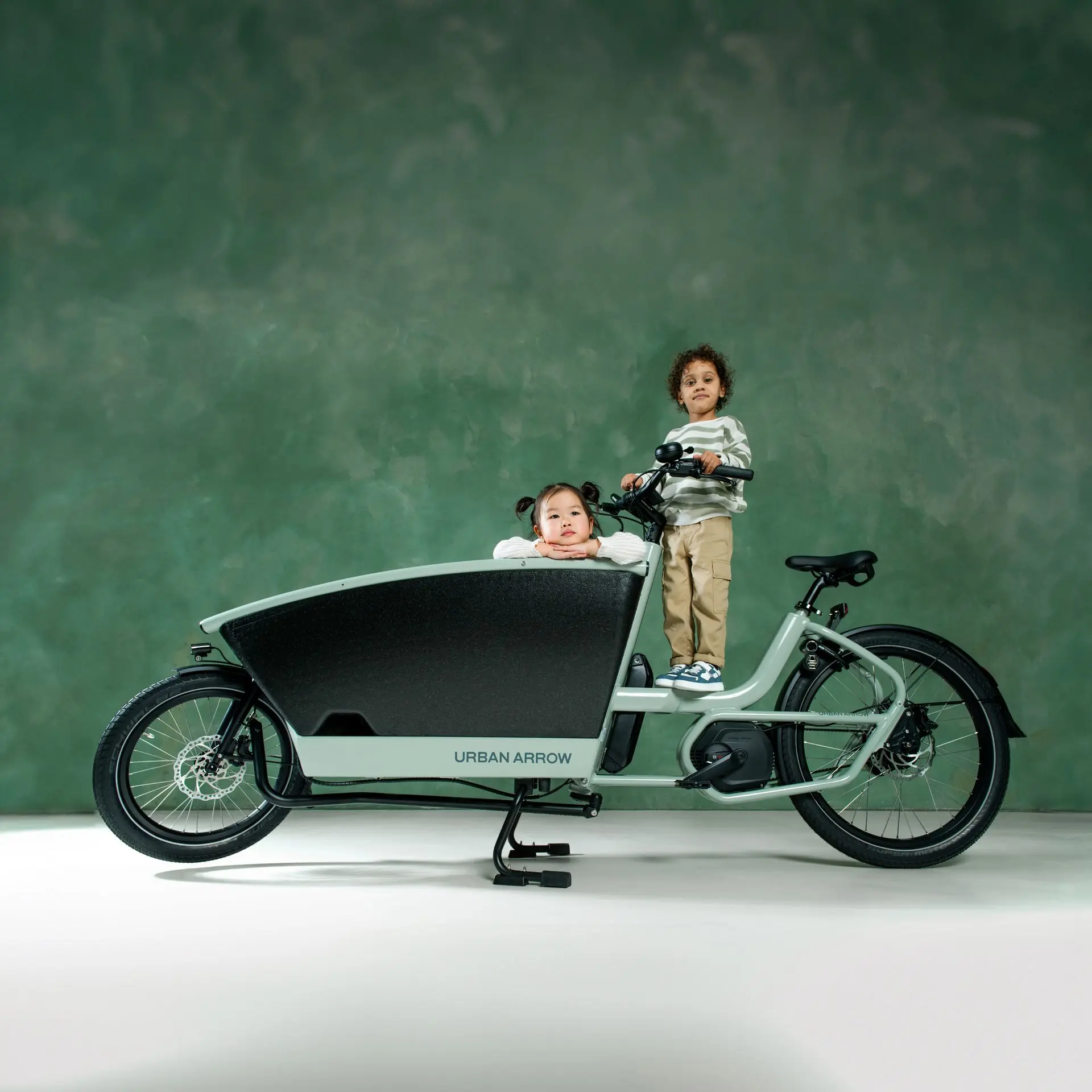 Entdecke das FamilyNext von Urban Arrow für Familien Zwei Kinder auf stehendem Lastenrad vor grüner Wand – spielerischer Moment und modernes Design vereint.