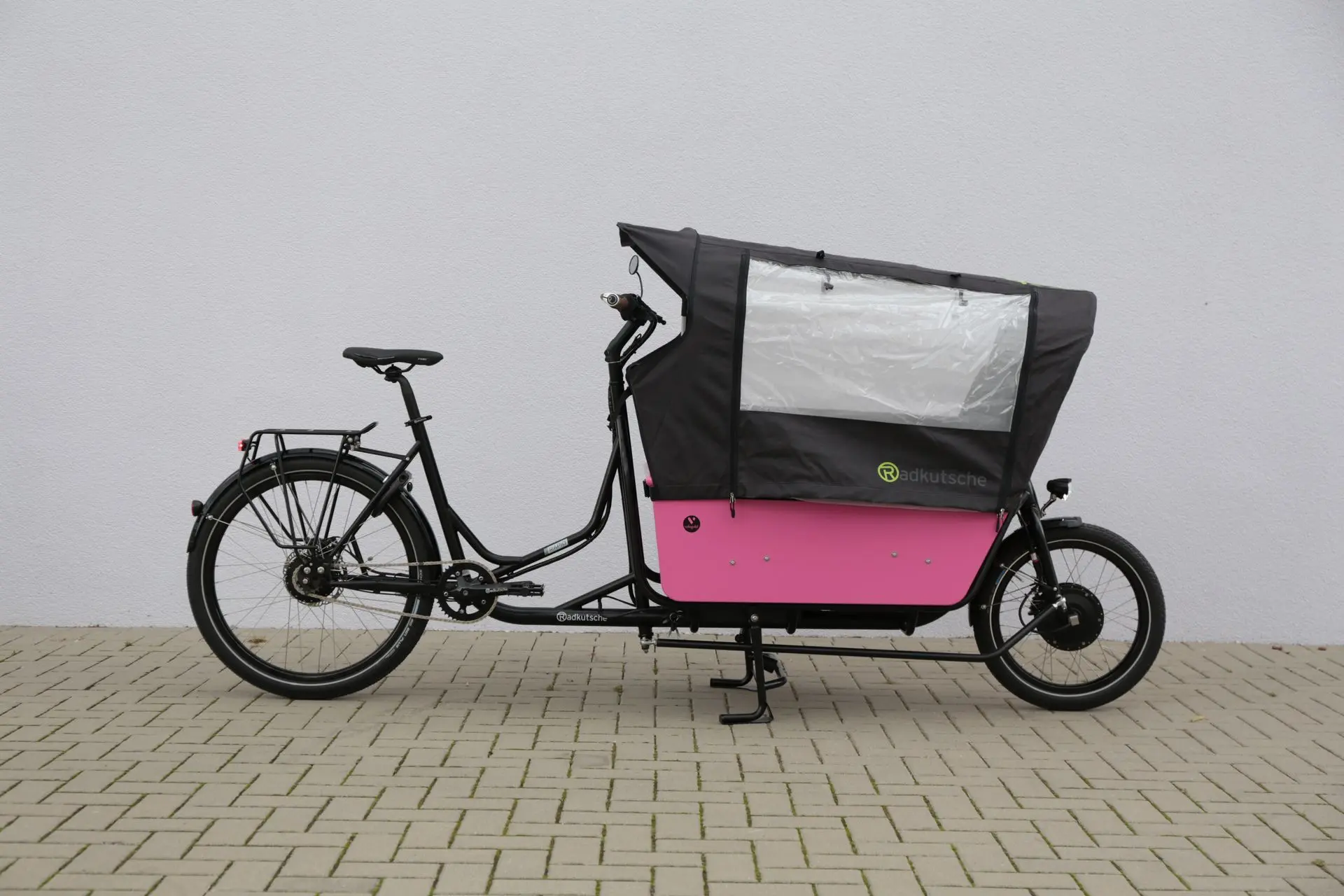 Radkutsche RAPID – das vielseitige Familien-Lastenrad mit Platz für Kinder & Gepäck. Radkutsche RAPID mit Transportwanne und Regenschutz-Zelt, abgestellt vor einer Hauswand – familienfreundliche Ausstattung.