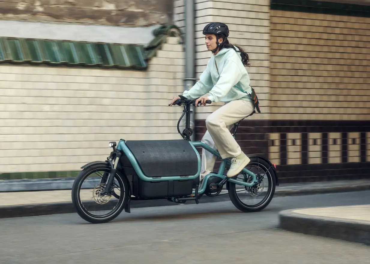 CARRIE von Riese & Müller – das smarte City-Lastenrad für deinen urbanen Alltag. Riese & Müller CARRIE – kompaktes Lastenrad mit geschlossener Box, gefahren von einer Frau entlang einer städtischen Hauswand.